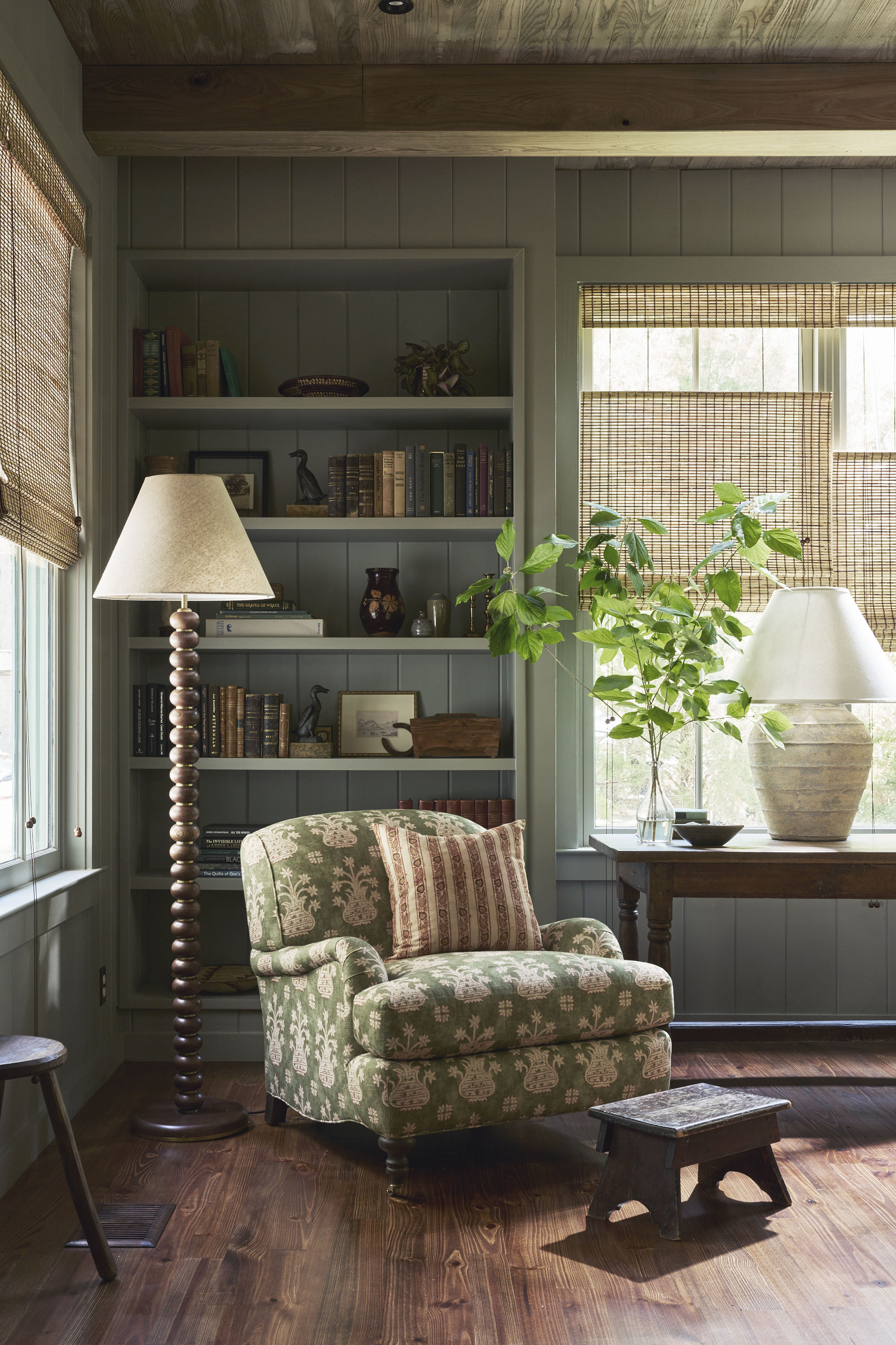 A reading corner in a mid-tone blue room with a built-in bookshelf, a bobbin floor lamp, and a green patterned, cozy reading chair. There are rattan blinds on the window.