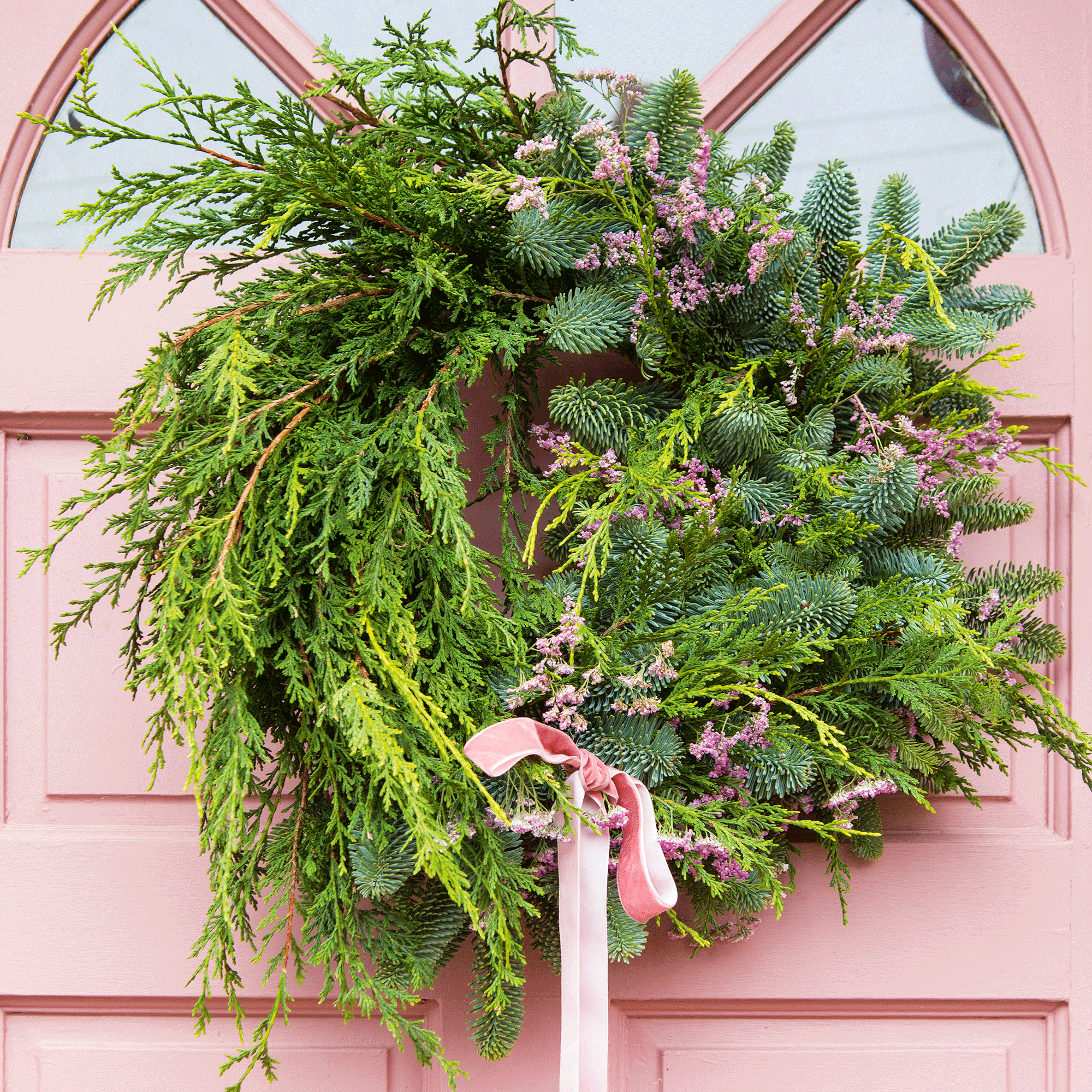 the front door of a house painted pink with a large foraged greenery wreath on the door tied with a pink bow