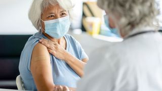 An older woman with white hair sits in a doctor's office with her sleeve rolled up to show a bandage on her arm, as if she'd received a vaccine. She and a doctor, seen in the foreground of the image, are both wearing surgical masks.