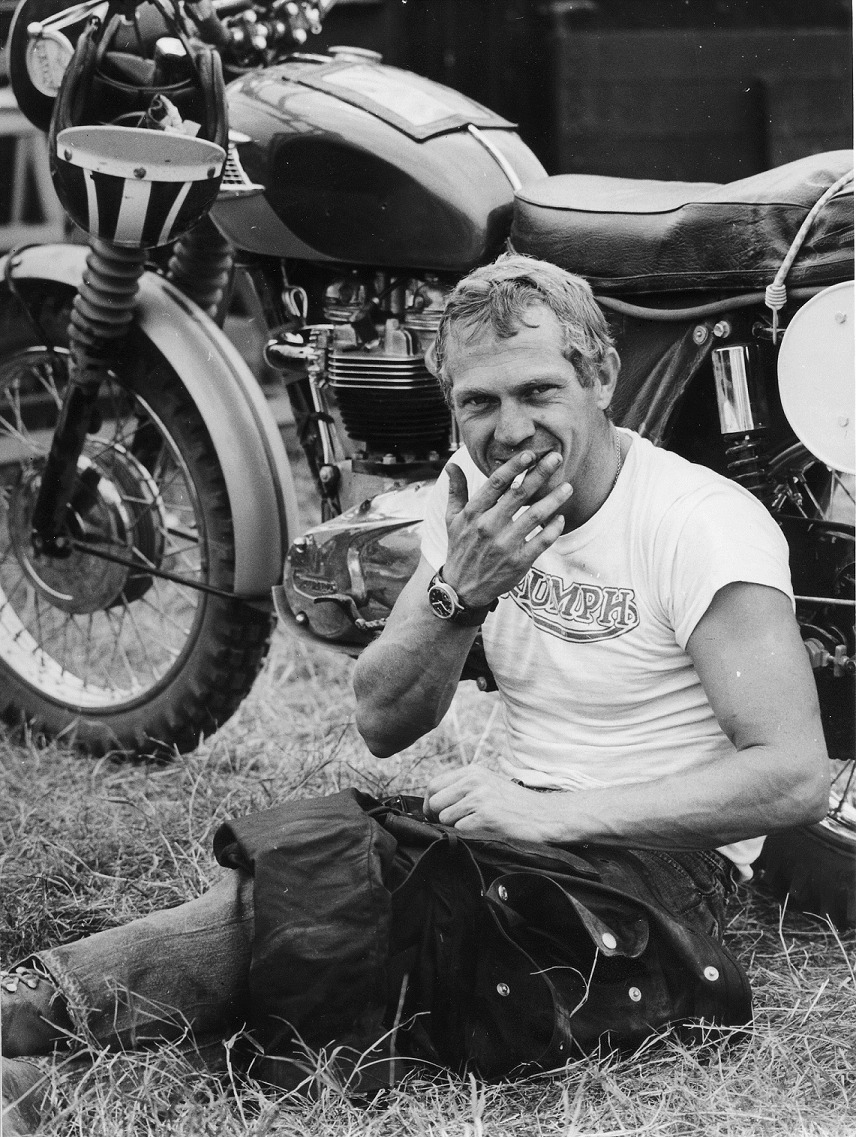 Steve McQueen sits on the grassy floor infront of his motorbike, dressed in a white Triumph T-shirt, smoking a cigarette 