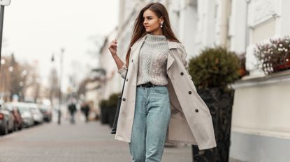 Model of a young beautiful woman in a fashion trench coat in a stylish knitted sweater with a leather black bag walks on the city near a white building. Elegant pretty trendy girl travels the street.