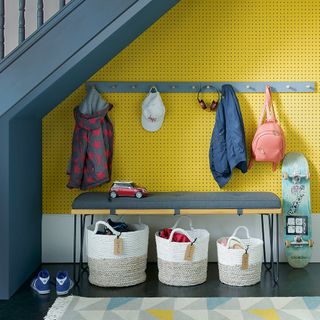 yellow peg wall with blue hanging hooks, underneath a blue staircase, with a blue bench seat and white storage baskets, and a skateboard leaning up against the wall