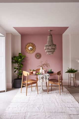 Pink color block painted wall section and ceiling with boho beaded chandelier, sleek round table and retro wood chairs, starburst wall mirrors, houseplants, and relaxed textiles.