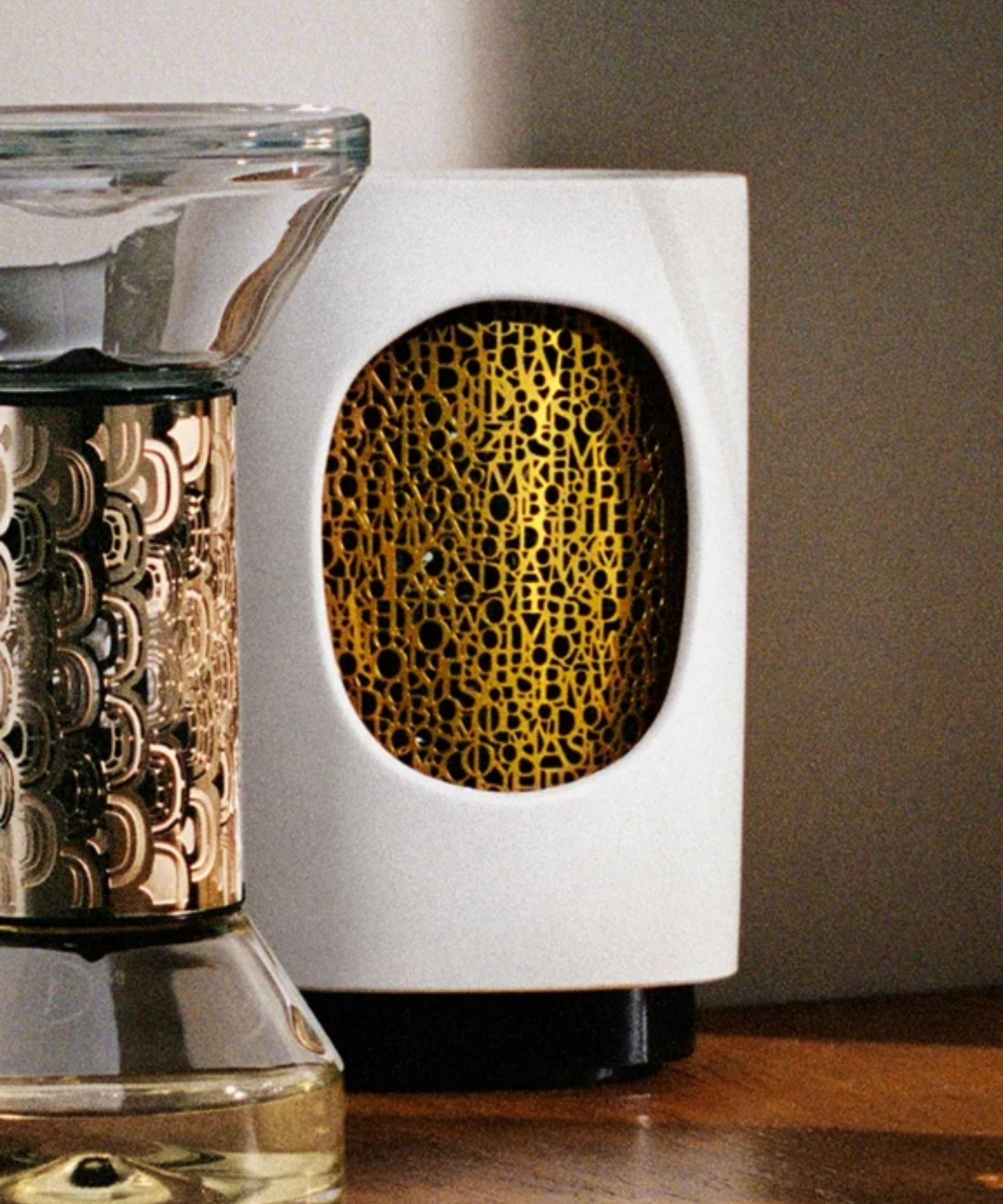 White Diptyque electric diffuser with gold detailing pictured alongside a glass hourglass diffuser