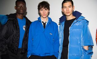 Three male models wearing looks from Off-White's collection. One model is wearing blue top, black trousers and black jacket with blue fur. Next to him is another model wearing a white shirt, black tie, black trousers and a blue jacket. And the third model is wearing a black top, black trousers and a blue padded coat with split sleeve design