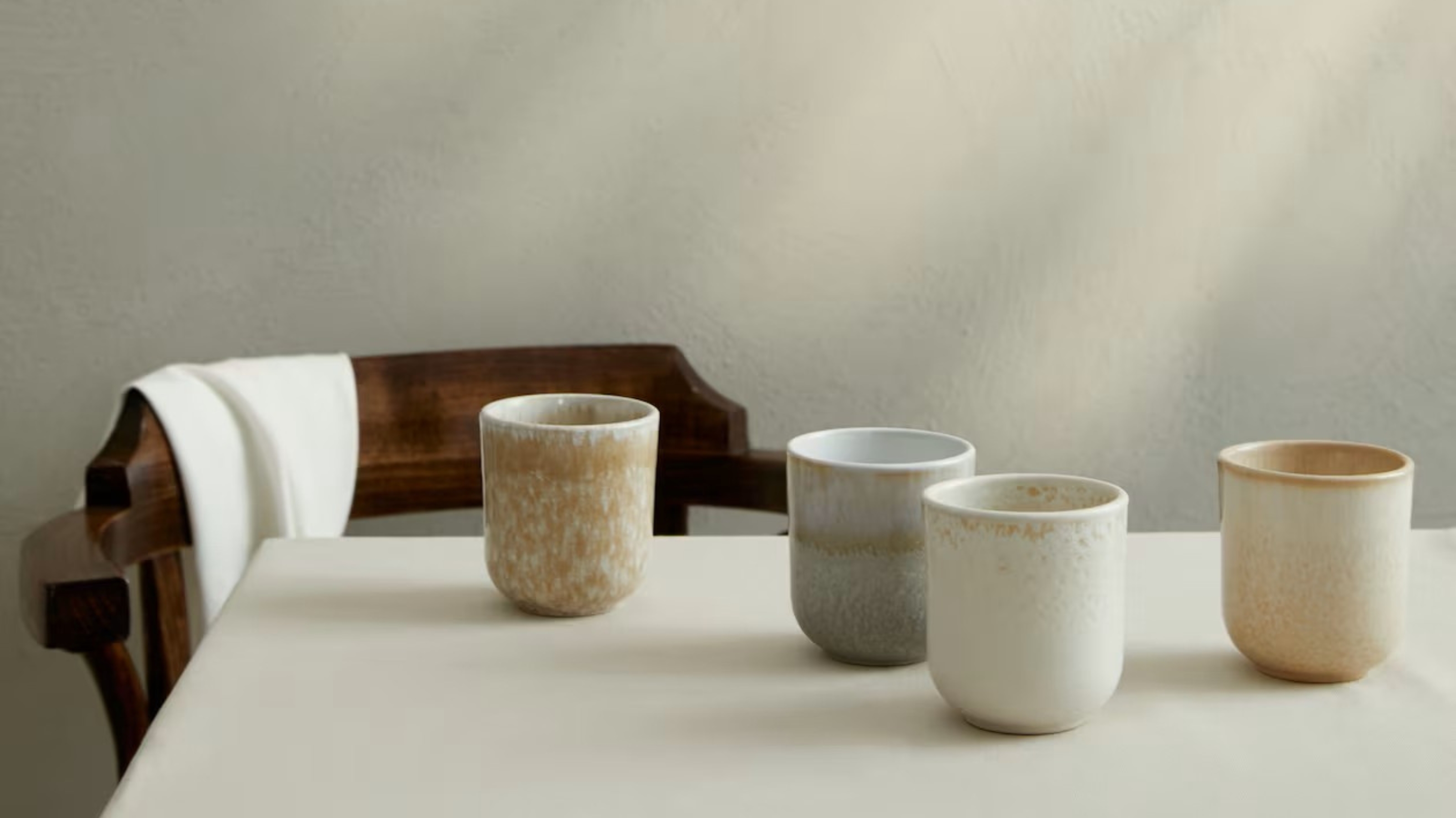 Image of the corner of a white table in a white room with a dark brown wooden chair pulled up to it. There are four ceramic, beige coffee mugs on the table.