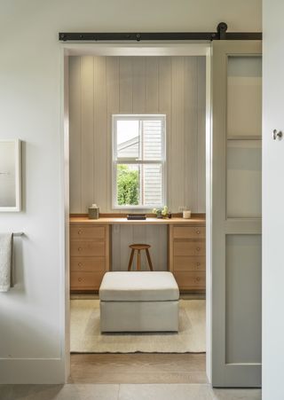 A modernly furnished study features a minimalist wooden desk and stool, a grey sofa, and a bright window surrounded by paneled wooden walls.