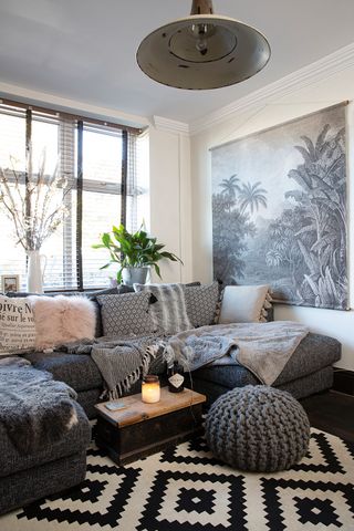 A small cosy living room with grey upholstered sofa and assortment of soft furnishings including faux fur cushions, throws, chunky knitted decor and palm tree wall hanging