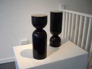 Two glosss black egg-timer shaped objects on a white box shaped surface, next to a white wall