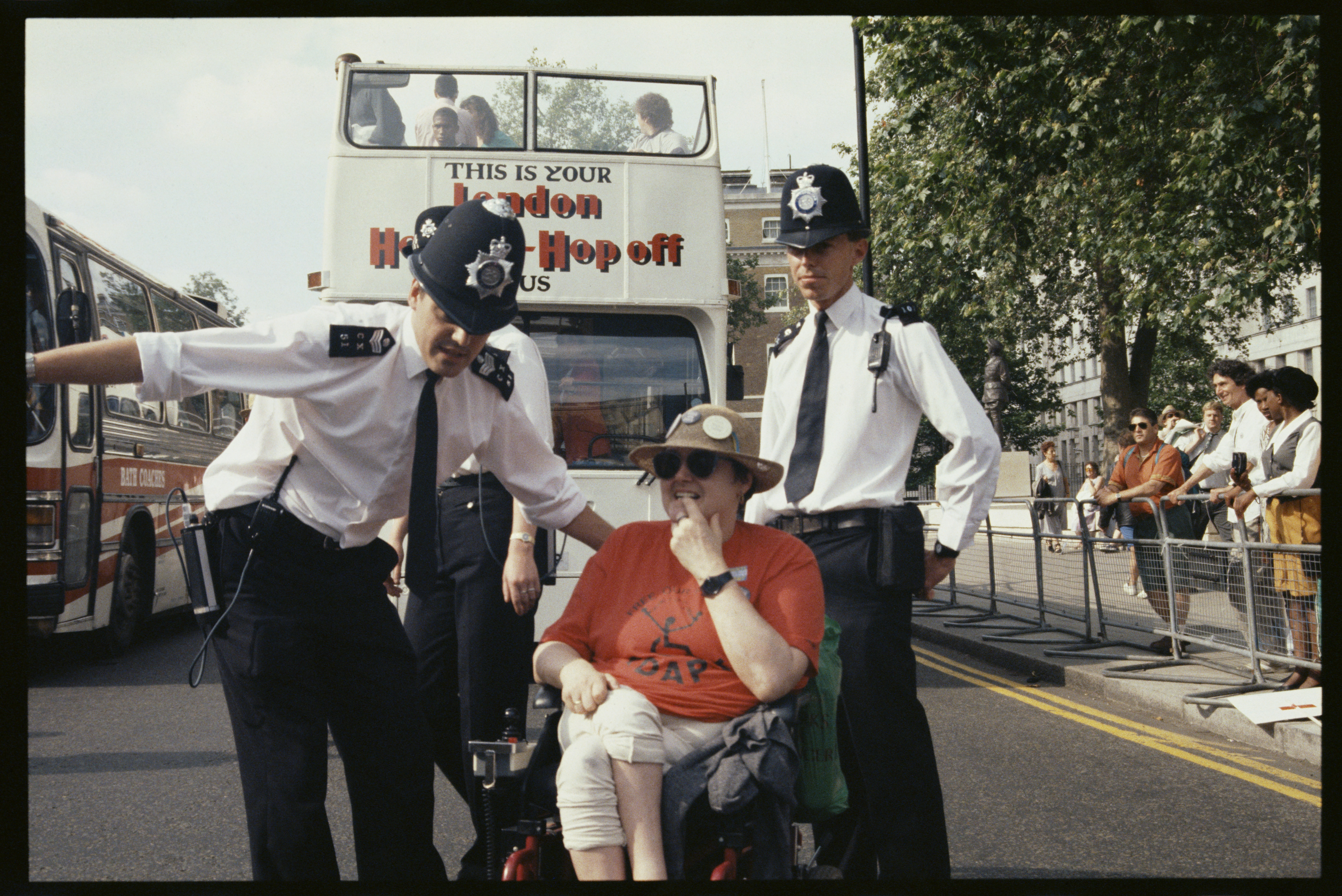 Two Metropolitan Police officers in shirtsleeves direct a wheelchair user in a red t-shirt and sun hat away from a road blocked by buses during a Whitehall disability rights protest.