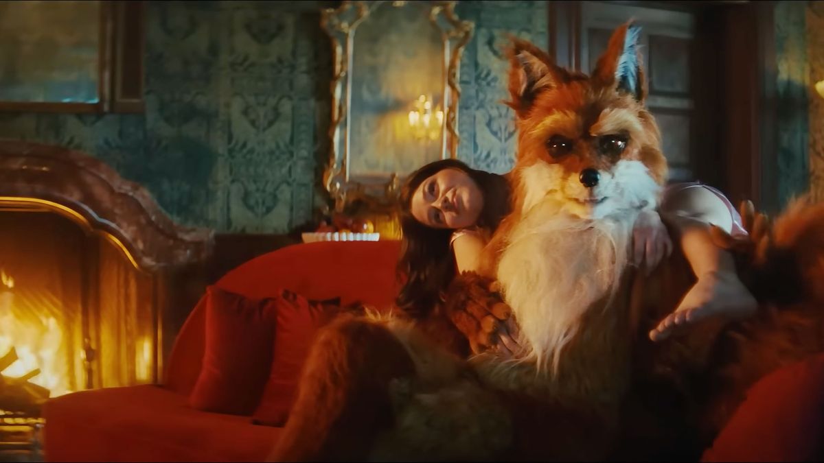 A shot of a humanoid fox in a dark room lounging on a sofa, with a human woman draped on its shoulders