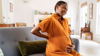 A heavily pregnant, African-American woman wearing and orange dress is sat on her couch holding her bump. She is grimacing as if she is starting labor