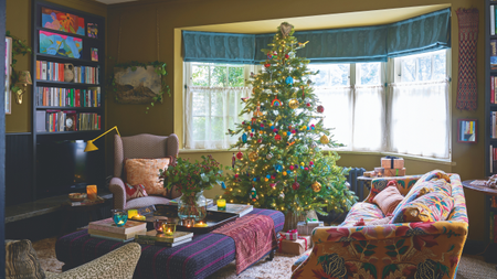 a nostalgic Christmas living room with a large tree decorated with colorful ornaments, a patterned couch, and an ottoman with a vase of flowers on top