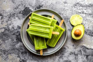 green avocado and lime ice lollies artfully arranged on a grey plate with a halved lime and avocado to the side for decoration