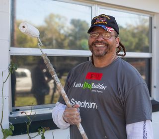 A man holds a paint roller wearing a Spectrum Community Assist shirt