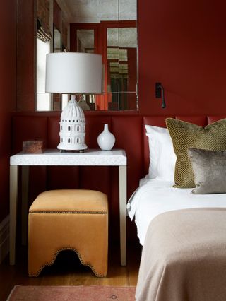 Close-up of bedroom with wall and padded headboard in burgundy, bed with white and olive green bedding, white marble bedside table and yellow-gold upholstered stool