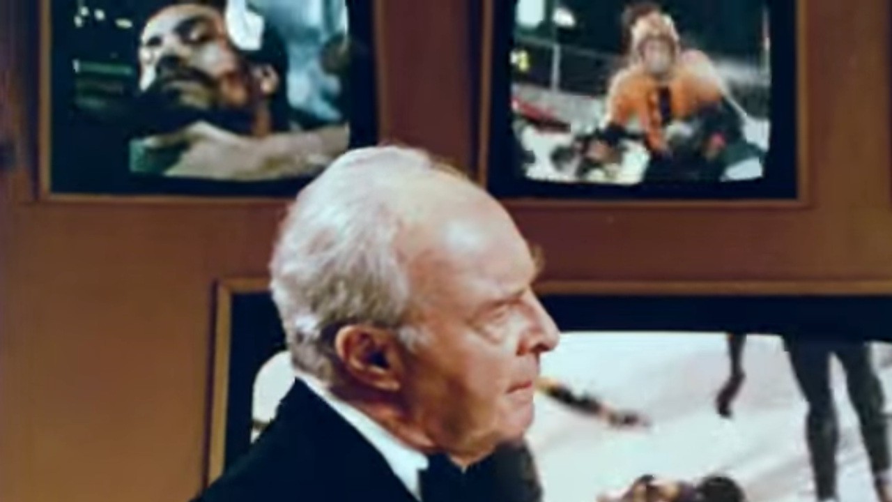 John Houseman in front of various screens in Rollerball