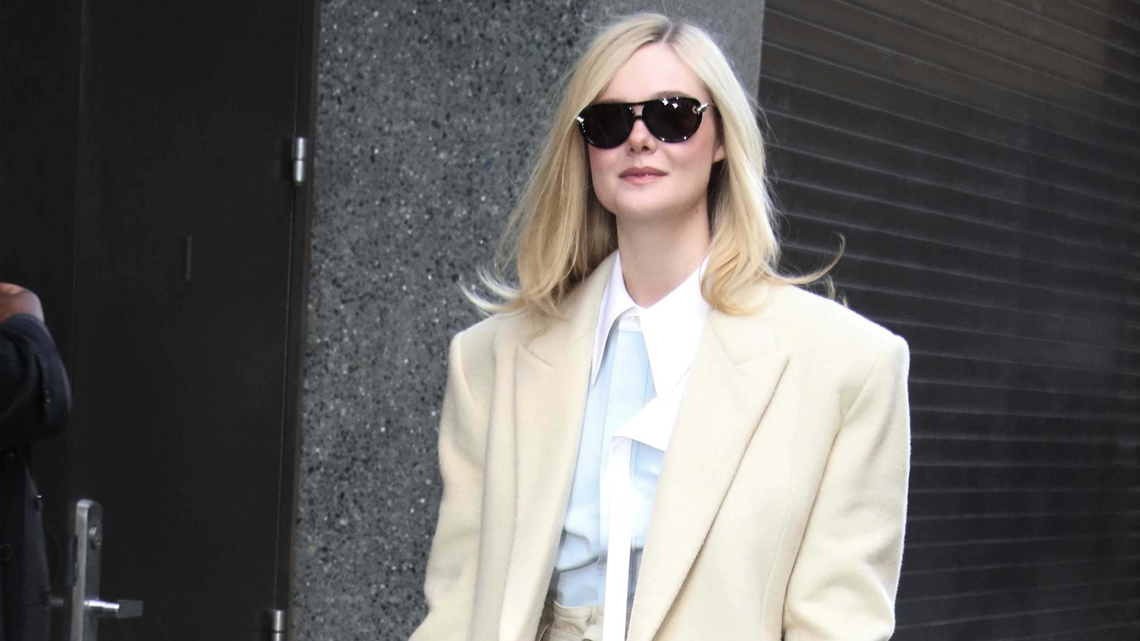 Elle Fanning wears a long ivory coat, white button-down, ivory trousers, ivory heels, and a black Bottega Veneta bag.