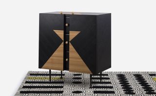 A black chest of drawers with light wood triangles on the front on an abstract black and white rug.