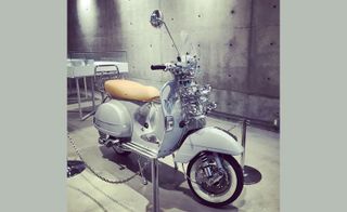 A white scooter with silver trim and a cream leather seat in front of a concrete wall.