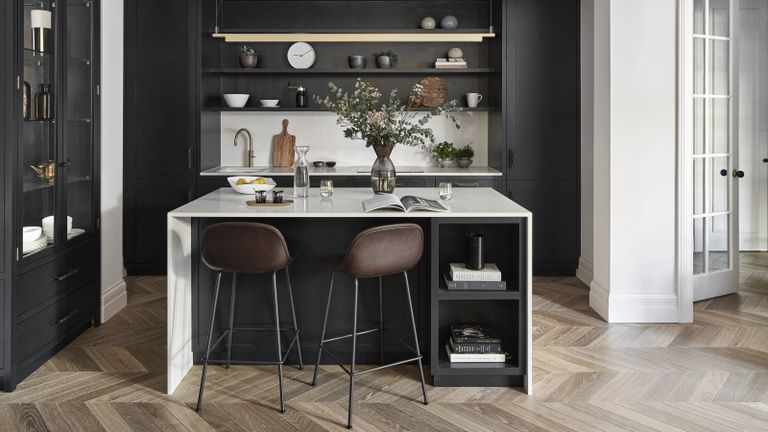 A kitchen with a black island with white wraparound counter top, seating and black floating shelves on the wall