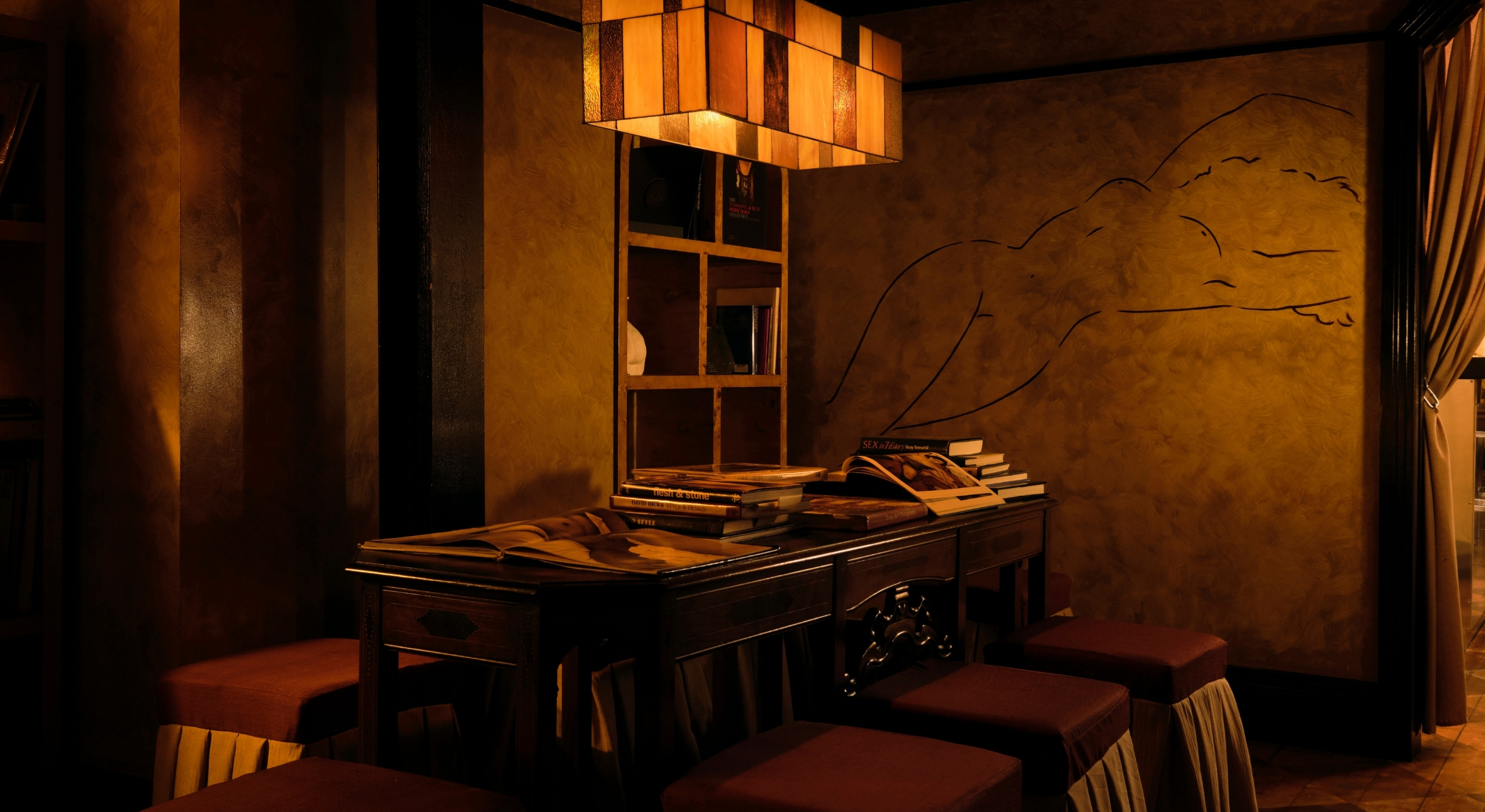 An atmospheric bar reception with a vintage wood desk, red leather and fabric stools, a pendant lamp in tempered squares of golden glass, and a mural depicting a woman stretching on her back nude.