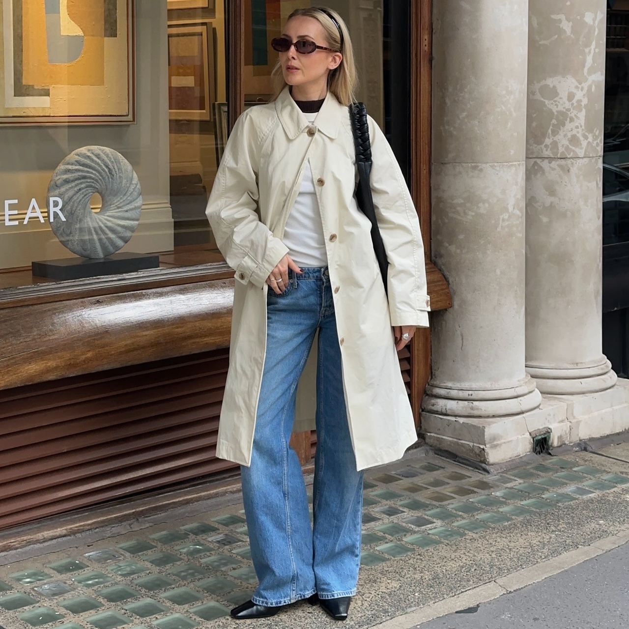 British style influencer Chloe Butler poses in London wearing a black headband, oval sunglasses, a black shoulder bag, an ivory trench coat, white top, relaxed jeans, and black boots