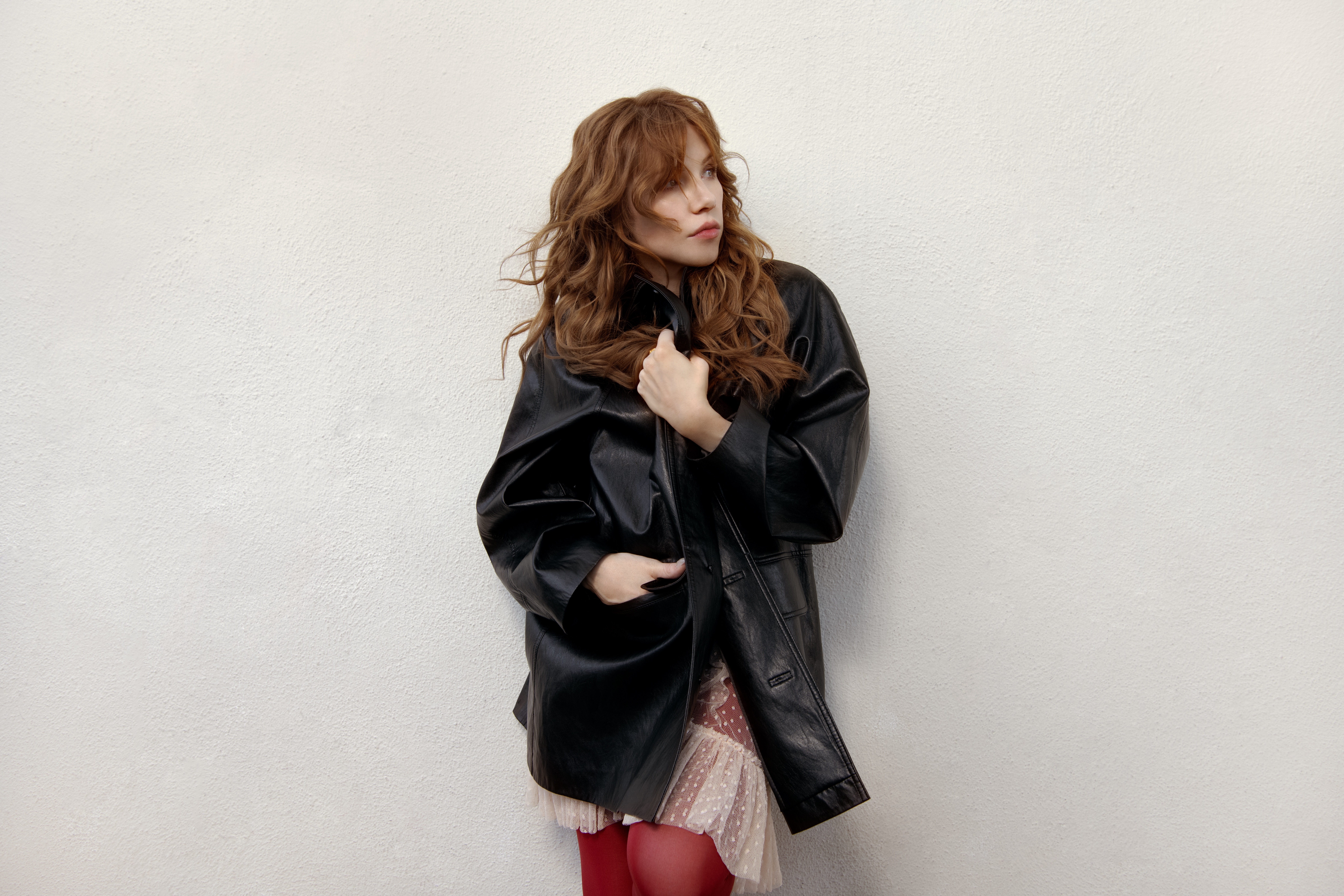 carly rae jepsen poses wearing a black oversized leather jacket and red tights against a white backdrop in a promo shot for day and night