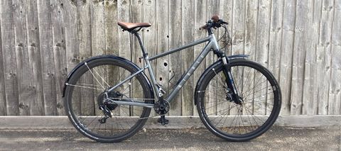 A grey Ribble Hybrid bike leans against a wooden fence