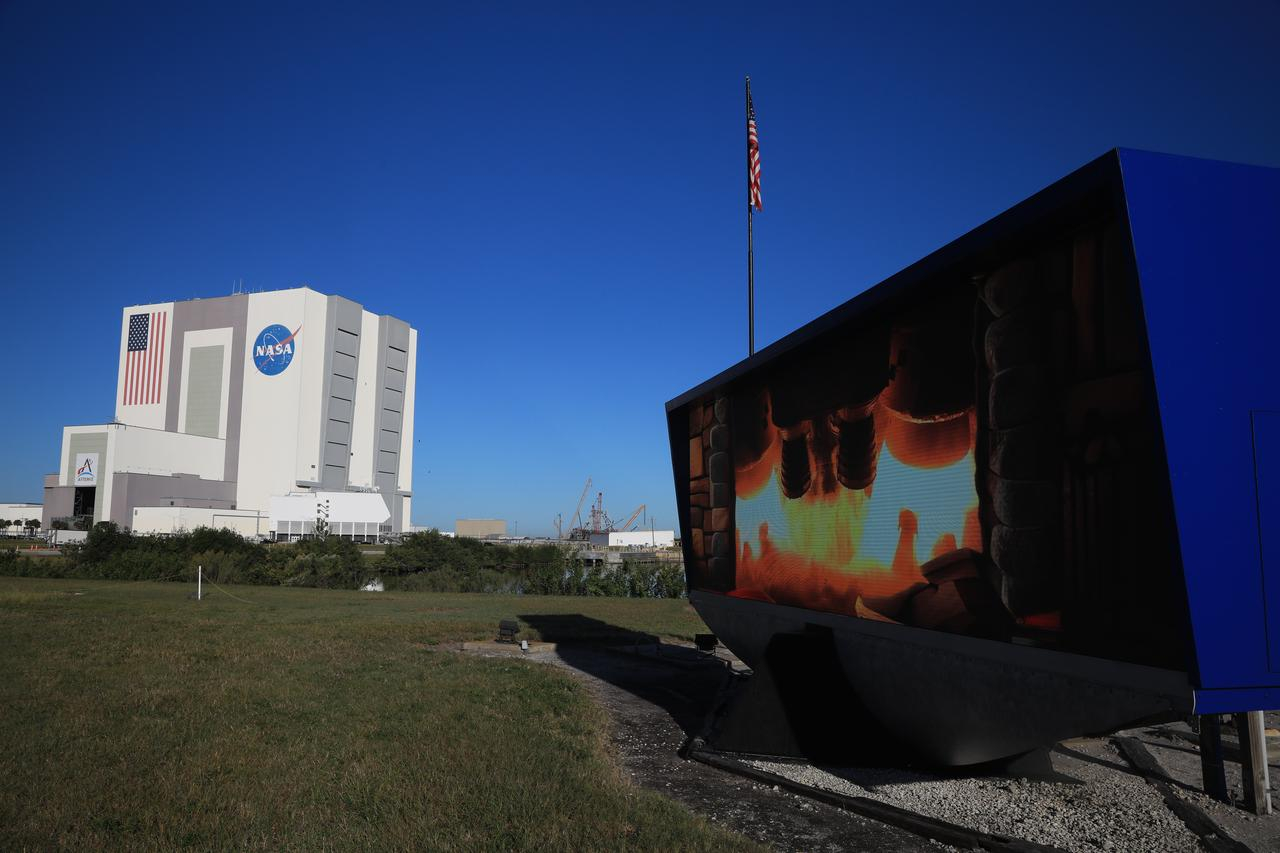 A grassy field leads to a large cube building stands against a blue sky in the background. In the foreground on the right, a blue screen showing the business end of a rocket as it launches.