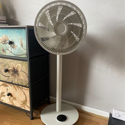 The Duux Whisper Flex 2 fan in pedestal mode on a wood-look floor next to a colourful chest of drawers in front of a taupe coloured wall