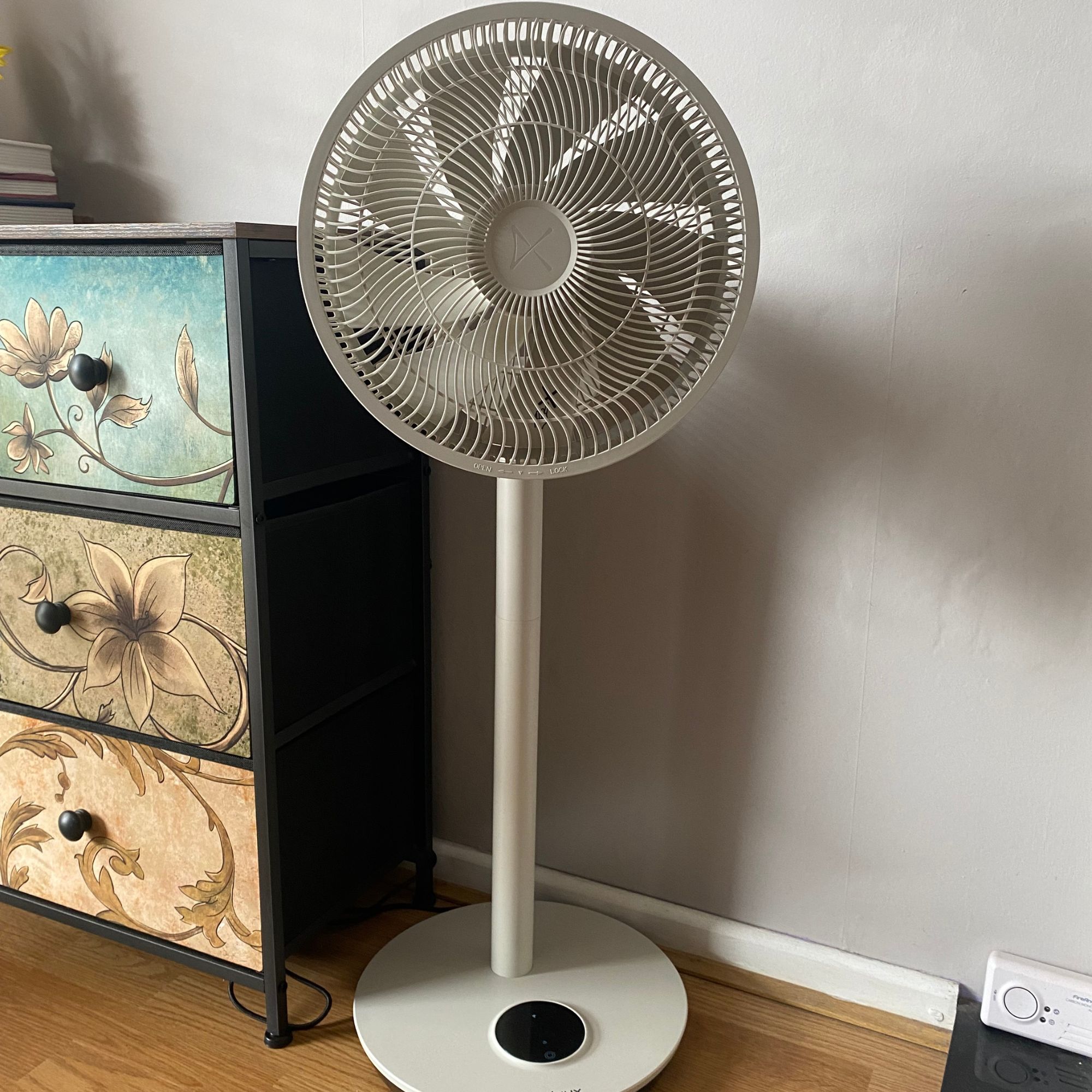 The Duux Whisper Flex 2 fan in pedestal mode on a wood-look floor next to a colourful chest of drawers in front of a taupe coloured wall 