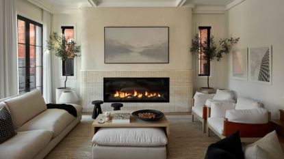 An off-white living room with potted trees, a long tiled fireplace, a pair of white armchairs, a wood coffee table, a pair of black stools, and a set of framed photos