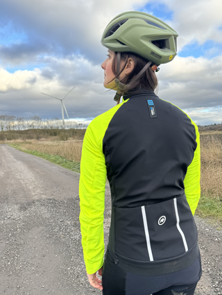 A woman looking away from the camera in a fluro yellow jacket and green helmet. The back of the jacket is black with large pockets and reflective detailing.