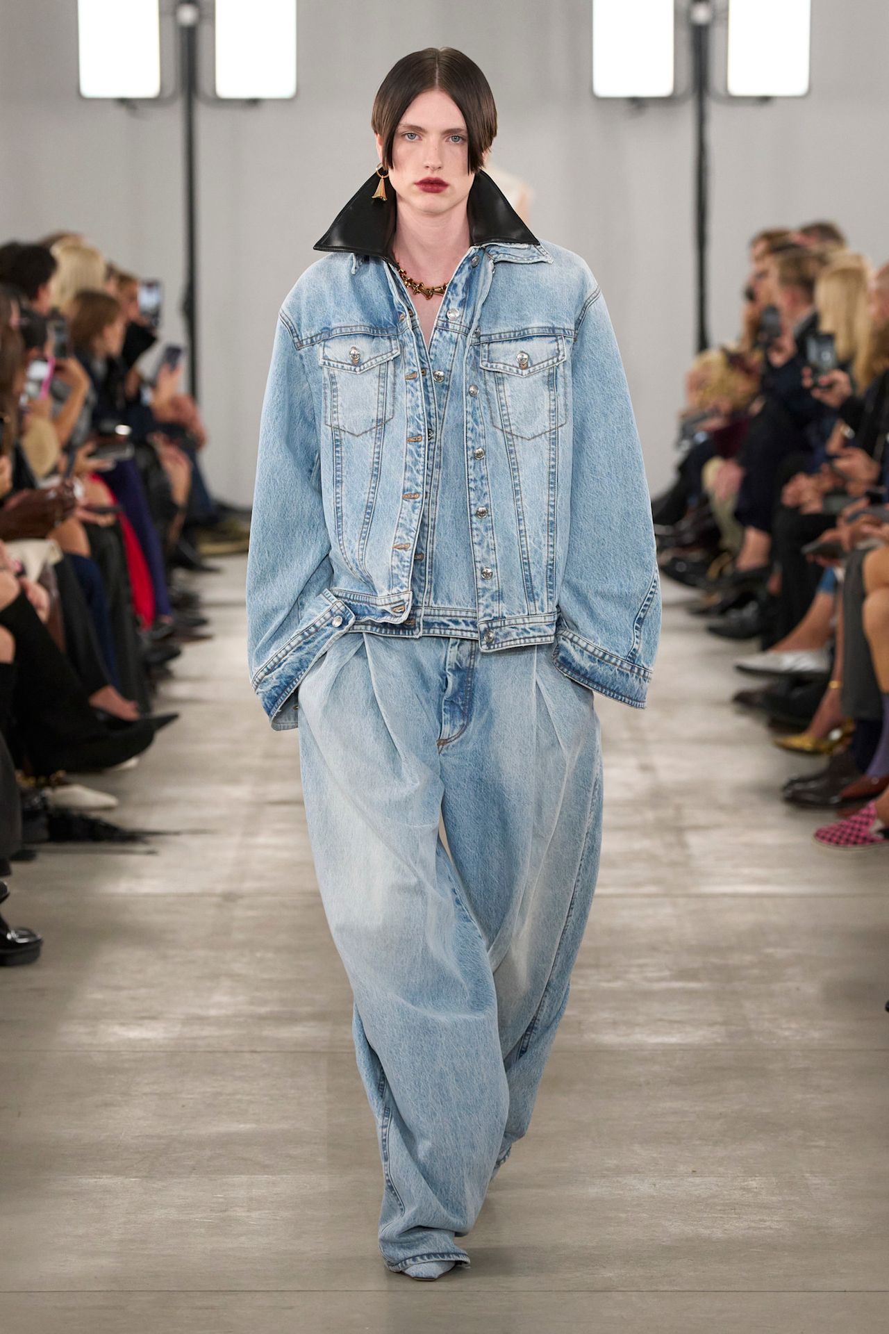 Layering staples for spring 2026 is shown in a photo from Sportmax's runway show, with a model walking while wearing an oversized denim jacket on top of another denim jacket that has a black leather collar styled with super baggy light wash jeans, denim-hued pointed pumps, a gemstone necklace, and a single gold earring