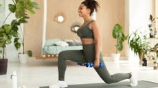 woman sideways to the camera, in khaki leggings and crop top holding a small blue dumbbell in each hand while doing a reverse lunge. there are plans in a living room setting blurred behind her.