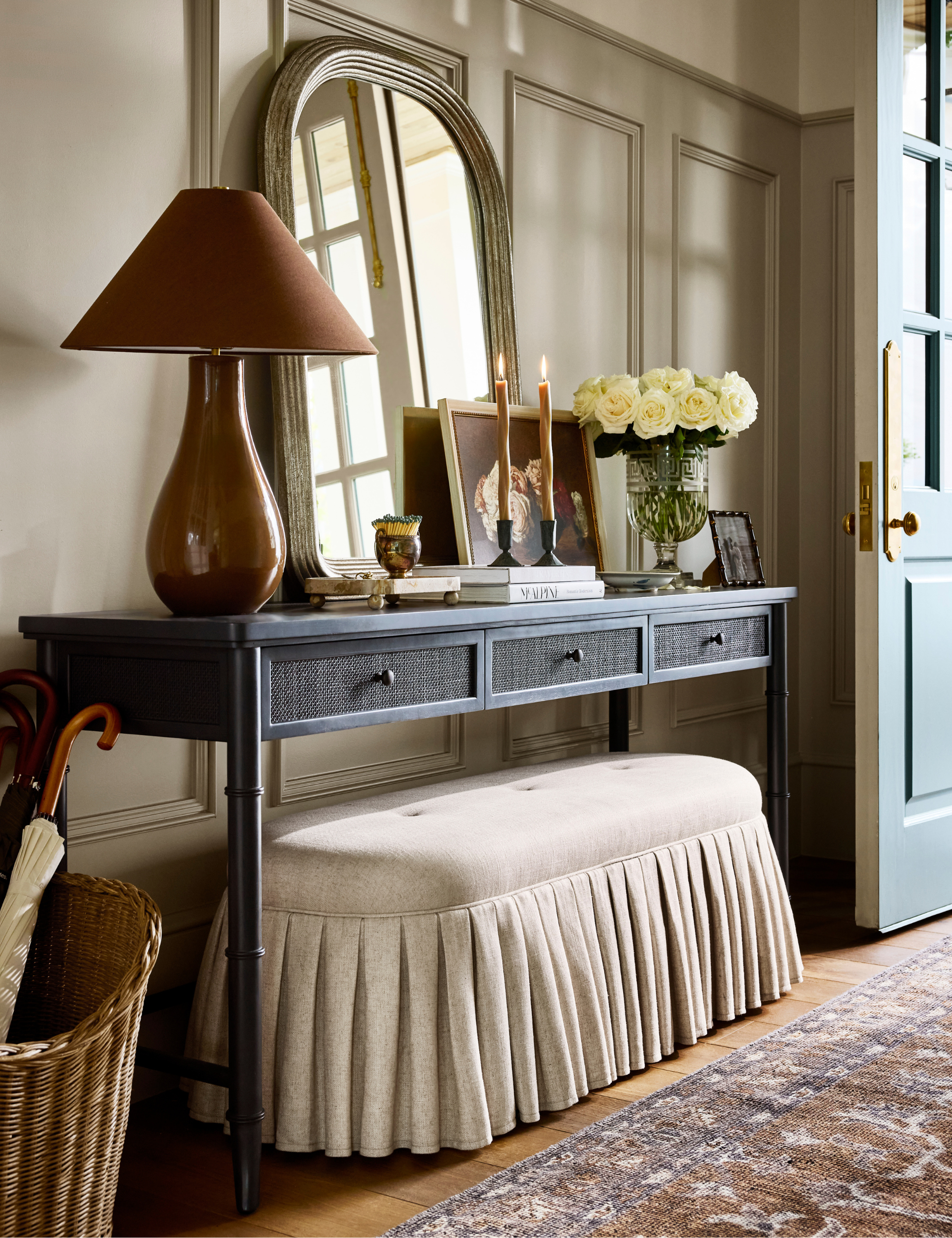 Richly layered entryway styled with a black wood-and-cane console table, a skirted oblong ottoman, an oversized table lamp, and a vintage rug