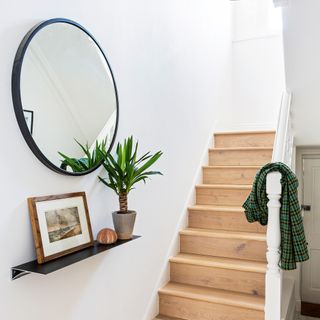 White hallway with wooden stairs with green jacket on railing with black framed circular mirror on wall