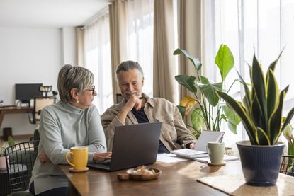A couple in their 60s discusses finances or other planning in front of a laptop. They sit at the dining room table with coffee, food and light coming through the windows.