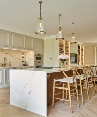 A kitchen with cream cabinets, a marble kitchen island, three wooden bar chairs, and three pendant lights on the white ceiling