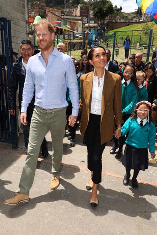 Prince Harry and Meghan Duchess of Sussex arrive at a school in Columbia the Duchess wears a brown blazer, white shirt and black trousers.