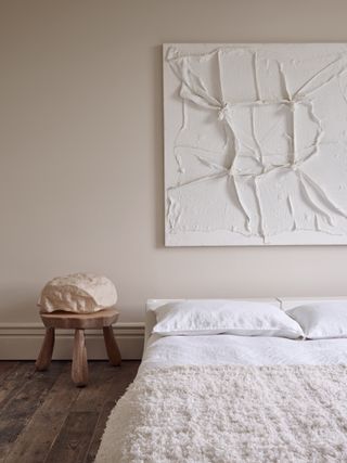 japandi bedroom with cream walls, white textured artwork hanging over bed, bed made with white sheets and a textured white throw, wooden stool as side table with stone decor styled on top