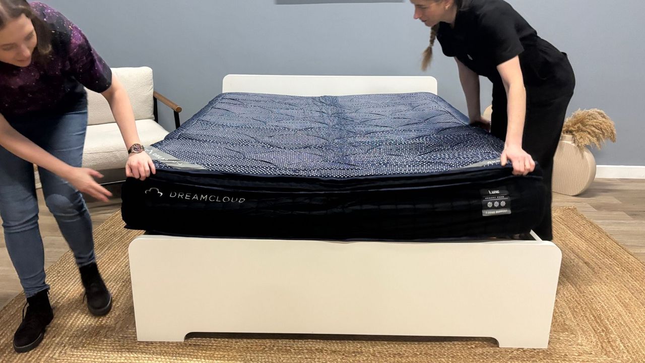 Two mattress testes unbox the DreamCloud Luxe Memory Foam mattress, taking it out of its box, cutting it out of its plastic wrapping and placing it on a white bed frame