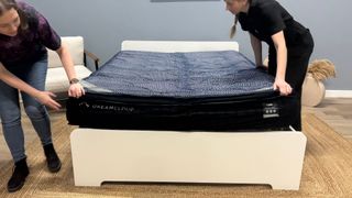 Two mattress testes unbox the DreamCloud Luxe Memory Foam mattress, taking it out of its box, cutting it out of its plastic wrapping and placing it on a white bed frame