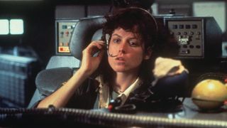 Ellen Ripley, a woman with medium length curly hair, is talking into a headset while sat on a desk chair with control panels around her