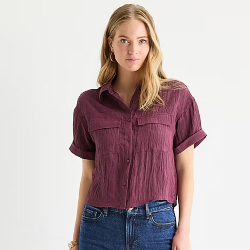 Blonde woman staring at camera, wearing indigo jeans and purple top with two pockets, white background. 