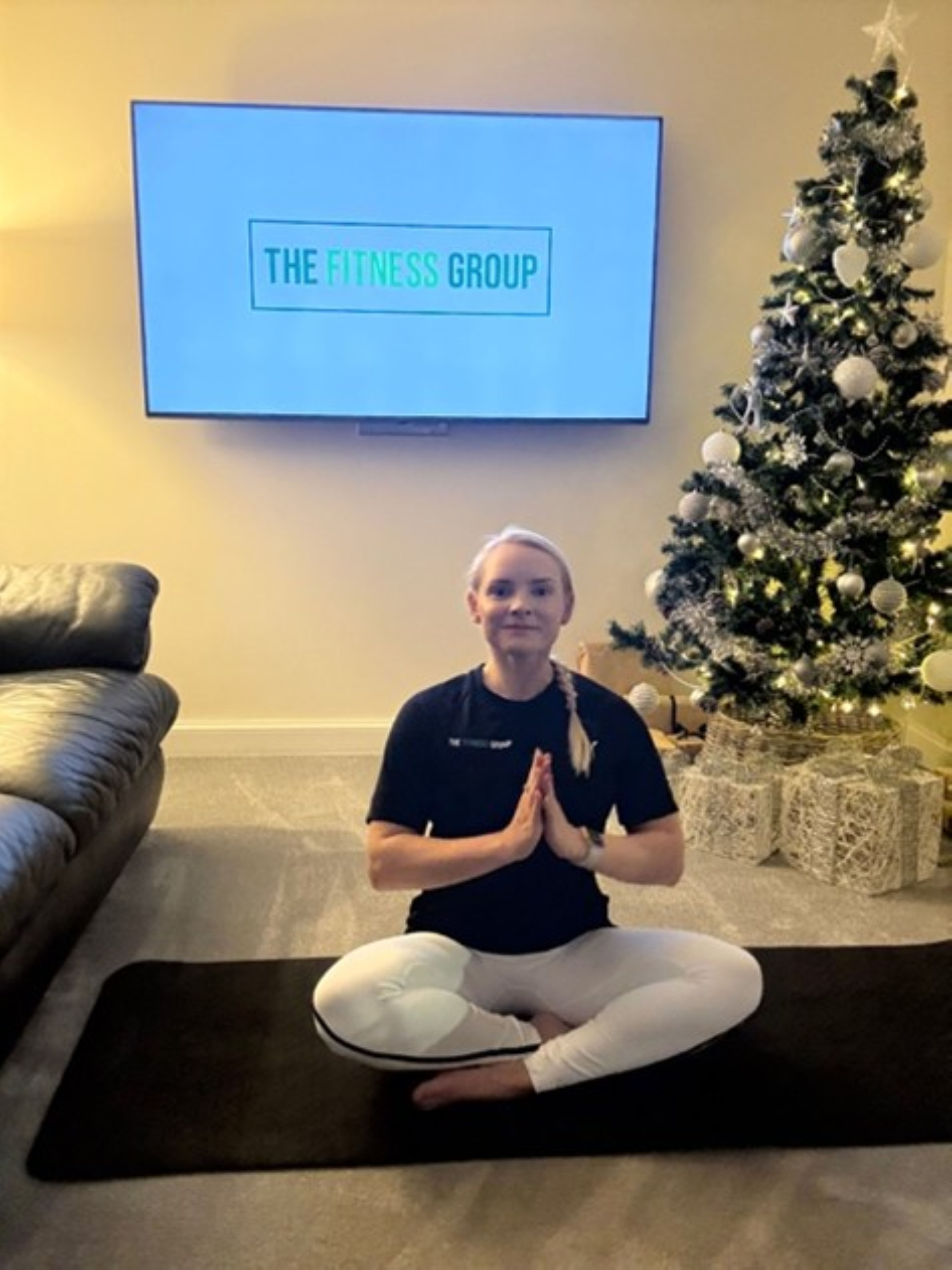 Woman demonstrates a Pilates exercise between a sofa and a Christmas tree. A mounted TV in the background reads "The Fitness Group"