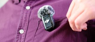 A photo of a DJI Mic 2 TX unit attached to the pocket of a burgundy shirt — the wearer of the shirt is adjusting the pocket for comfort. The TX unit is fitted with a wind shield.