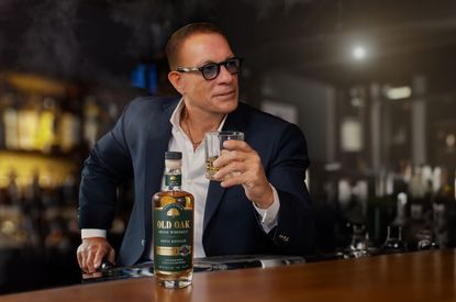Jean-Claude Van Damme sitting at a bar with a glass of Old Oak whiskey in his hand and the bottle on the bar