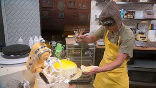 A man wearing a hood and goggles airbrushes a yellow cake (partly hidden behind a stand mixer) in an industrial kitchen, in 'Nailed It!'